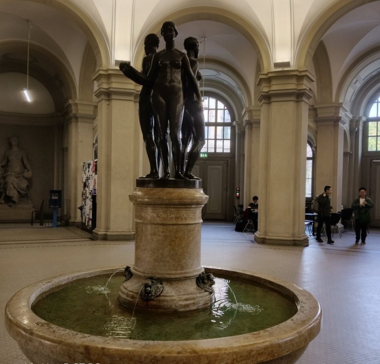 Fontanna Trzech Gracji w głównym holu ETH Zürich. Trzy kobiece postacie z brązu wieńczą kamienny postument rzeźby Eduarda Zimmermanna z 1921 roku.”